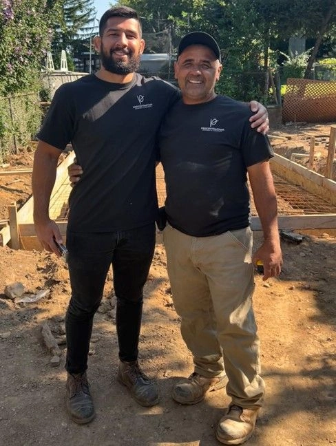 A father and son in professional contractor uniforms smiling at a construction site, symbolizing family-run business values.
