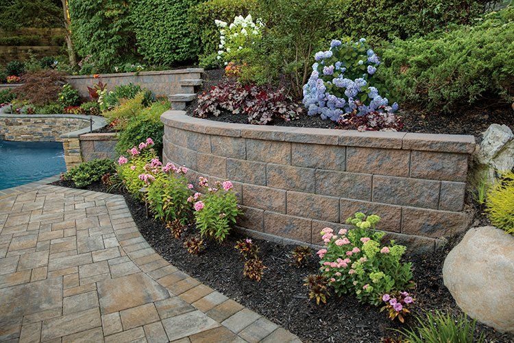 A sturdy concrete retaining wall in a residential garden, supporting tiered landscaping with various plants and paved walkways, demonstrating functional and aesthetic exterior design.